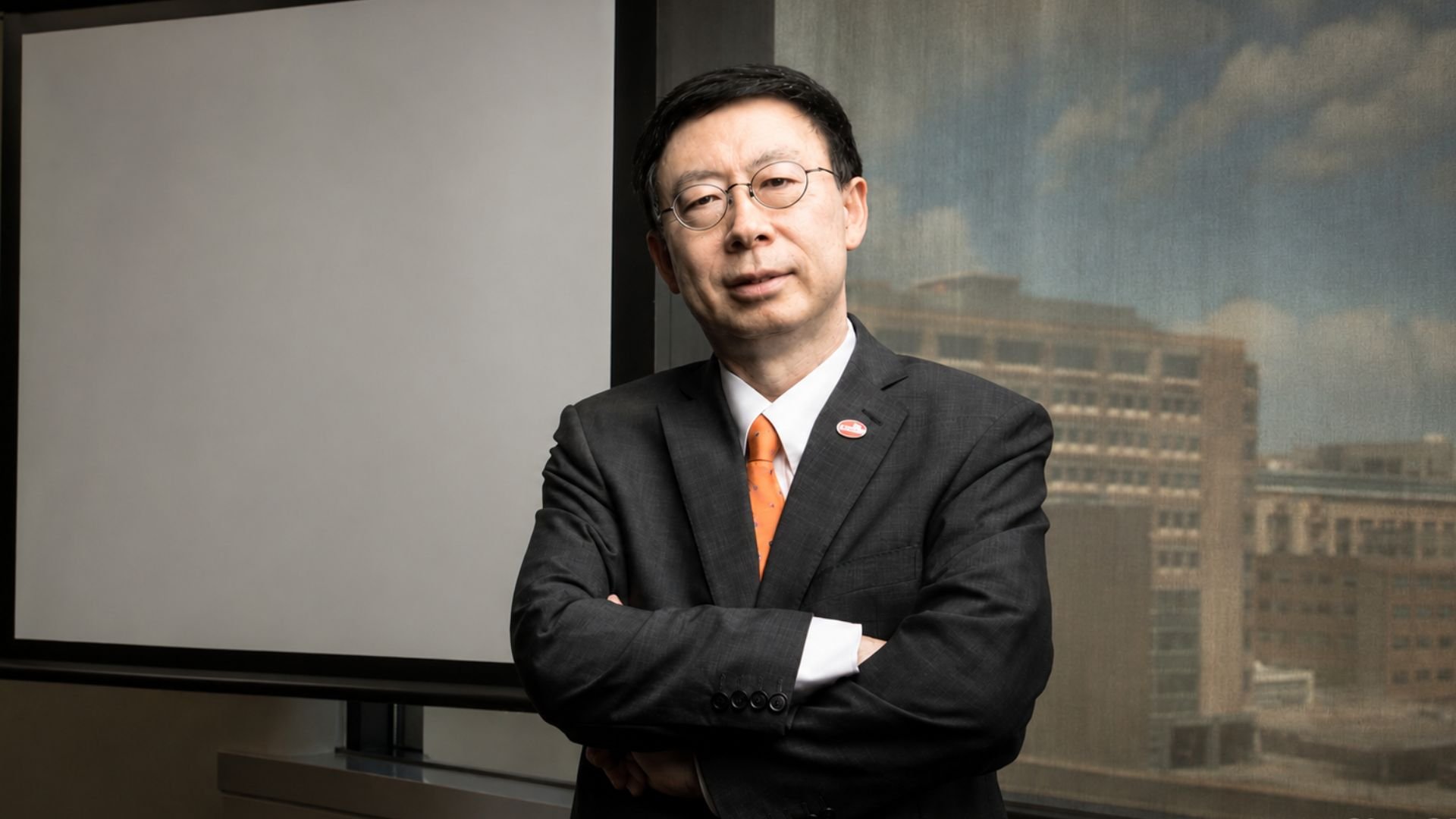Person in a dark suit and orange tie stands with arms crossed in front of a window overlooking city buildings and a projection screen.