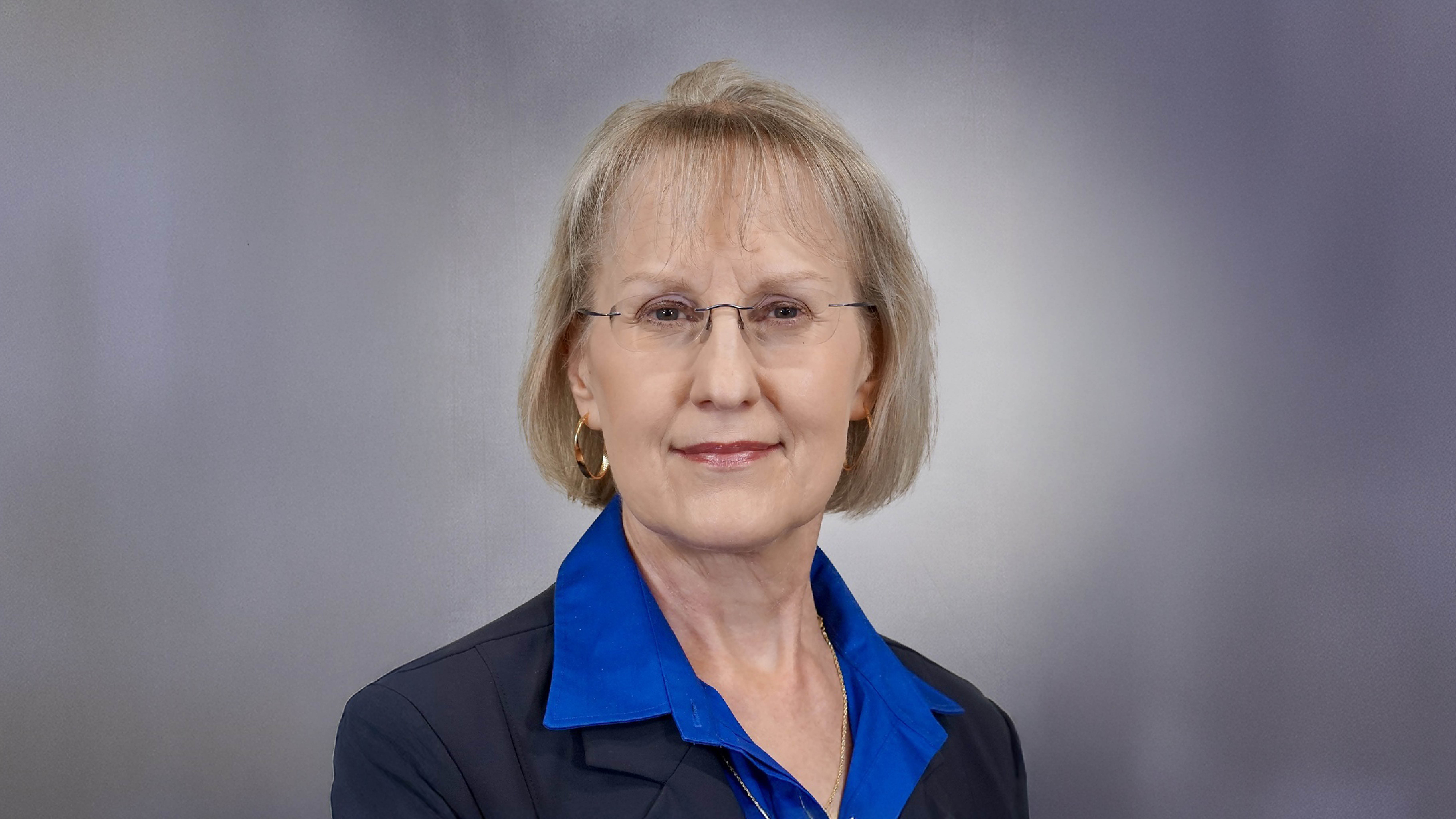 Professional headshot of a person wearing glasses, a blue collared shirt, and a dark blazer, posed against a gray gradient background.