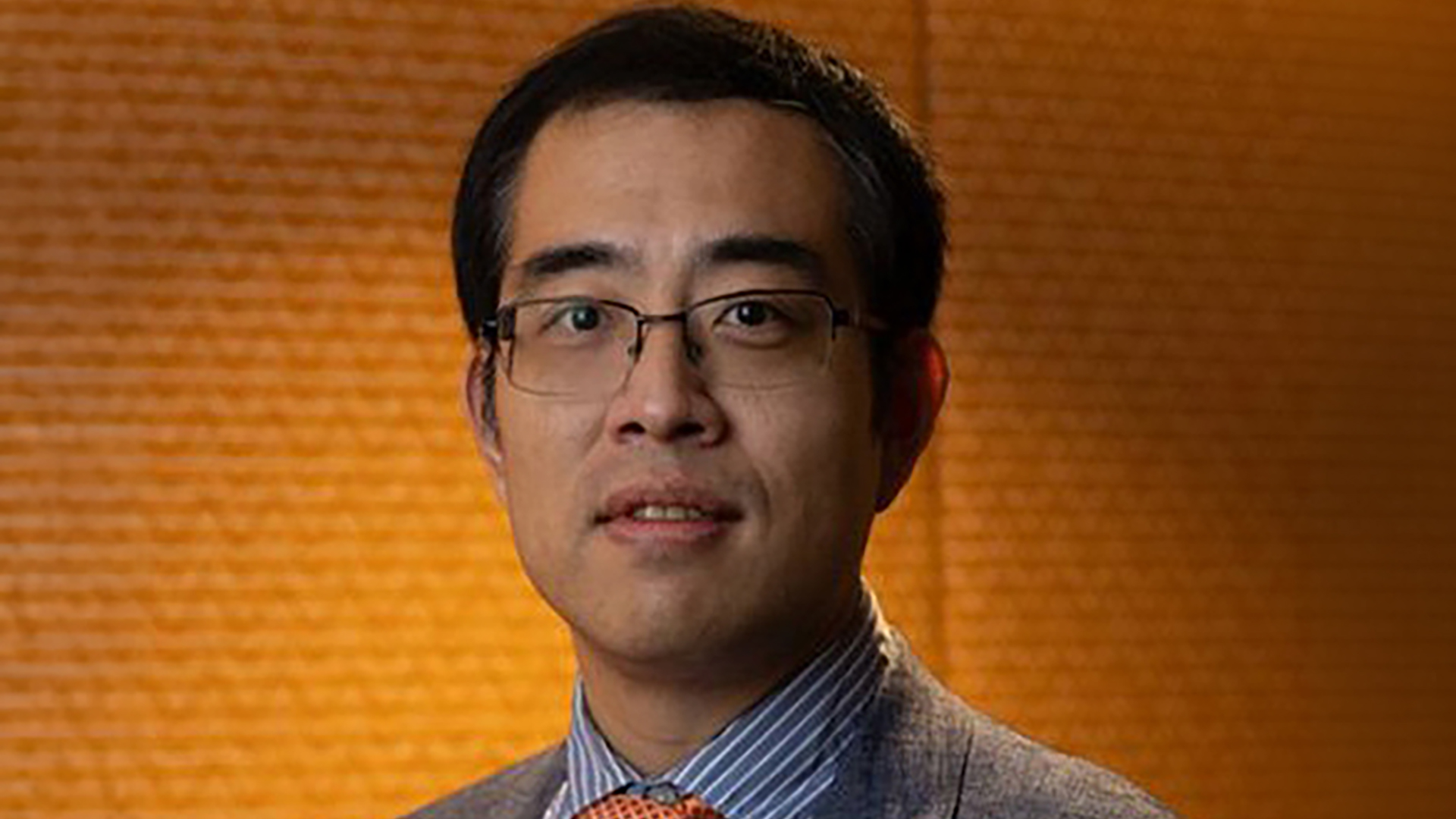 Professional headshot of a man wearing rectangular glasses, a gray suit jacket, striped dress shirt, and patterned tie, posed against a warm orange-brown textured background.