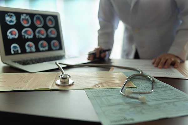 Photo of a screen of brain images and a physician in the background and a stethoscope in the foreground. Photo credit is Getty Images.