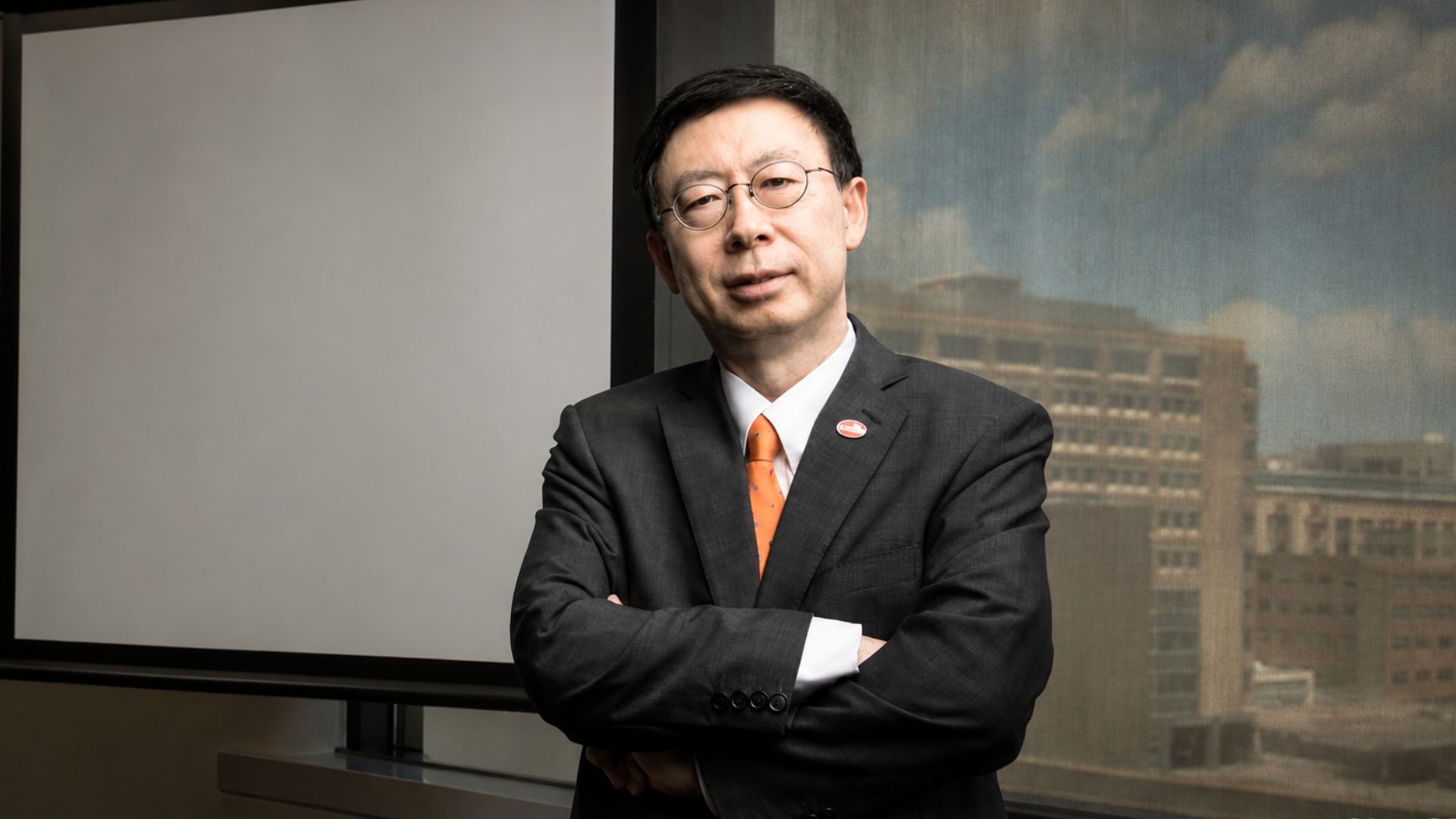 A professionally dressed man stands with arms crossed in a modern office, wearing a dark suit and orange tie. He is positioned beside a blank projection screen, with a large window behind him showing a blurred cityscape.