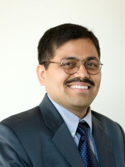 Sameer Bhat, in coat and tie, smiling, wearing glasses